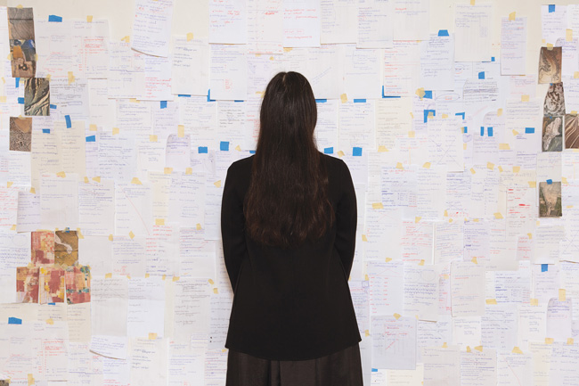 the artists with their back facing the camera. wall covered with letter size documents