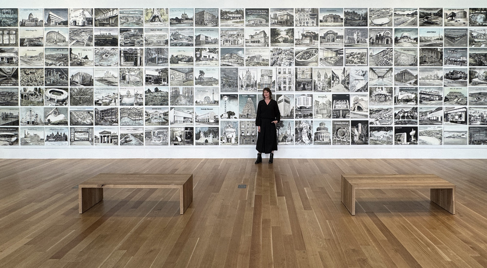 artist Ellen Harvey stanging in a gallery in front of a wall filled with hundreds of small drawings